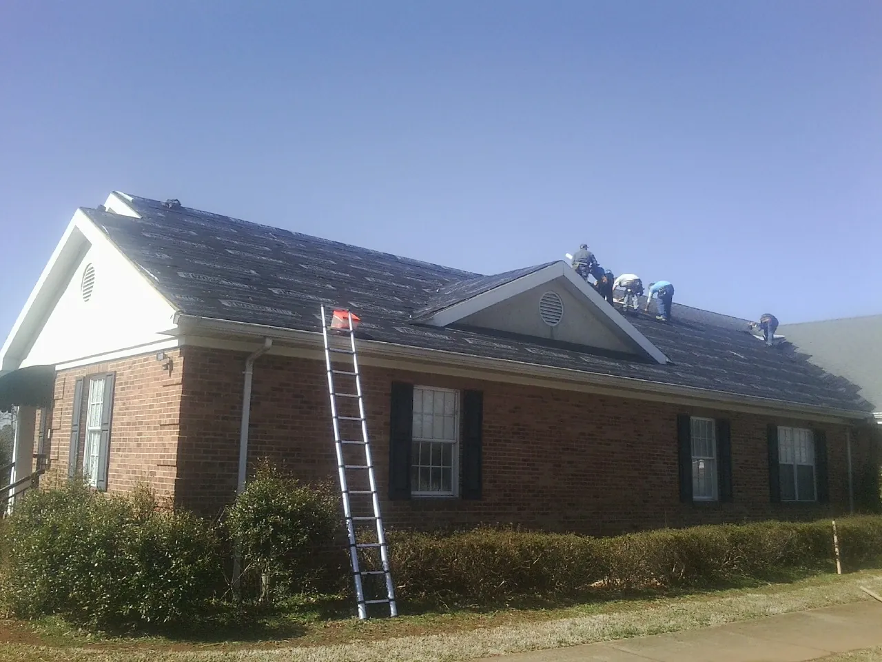 Crew tearing shingles off a brick ranch roof with a ladder set — Emergency Roof Repair work by our crew in Shabbona