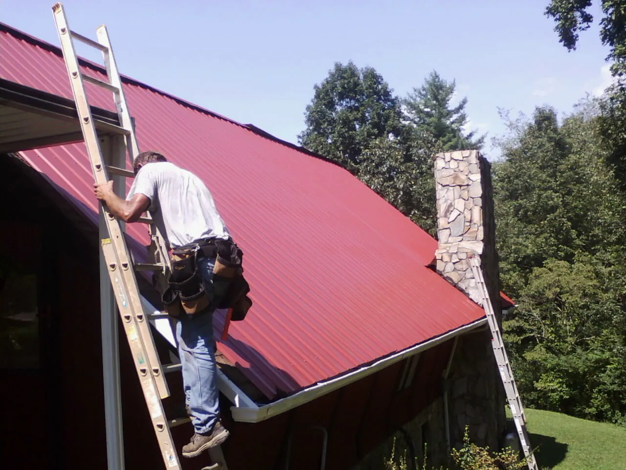 Roofer on a ladder beside a red metal roof and stone chimney — Soffit & Fascia Repair by Roofing Brothers in Shabbona